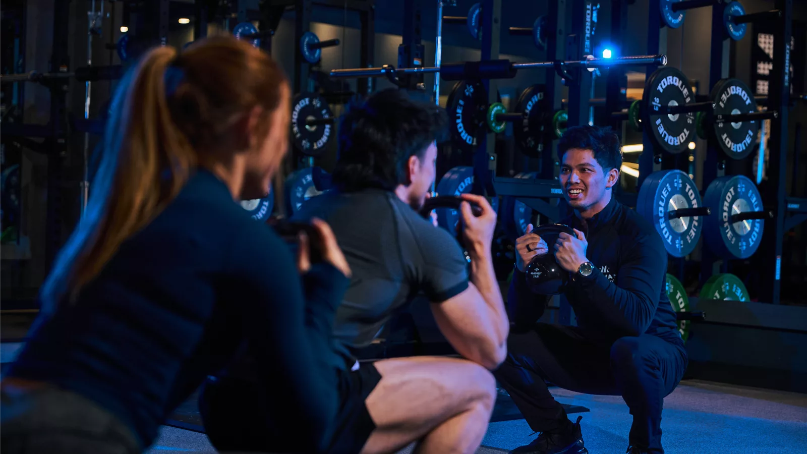 People engaging in group personal training, guided by a trainer in a gym setting with weights in the background.