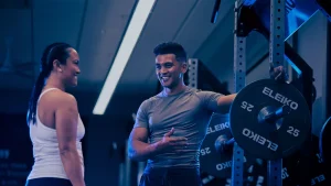 A trainer and a beginner engage in group personal training at a gym, discussing workout techniques by a weight rack.
