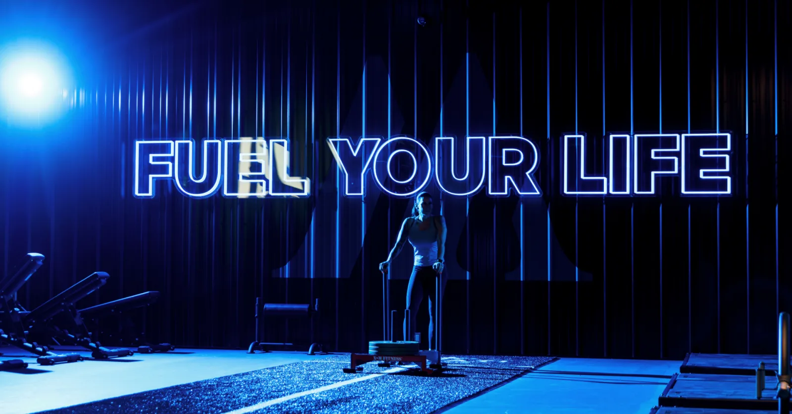 Person in gym under neon sign "FUEL YOUR LIFE," emphasizing HYROX Recovery focus in a dynamic training environment.