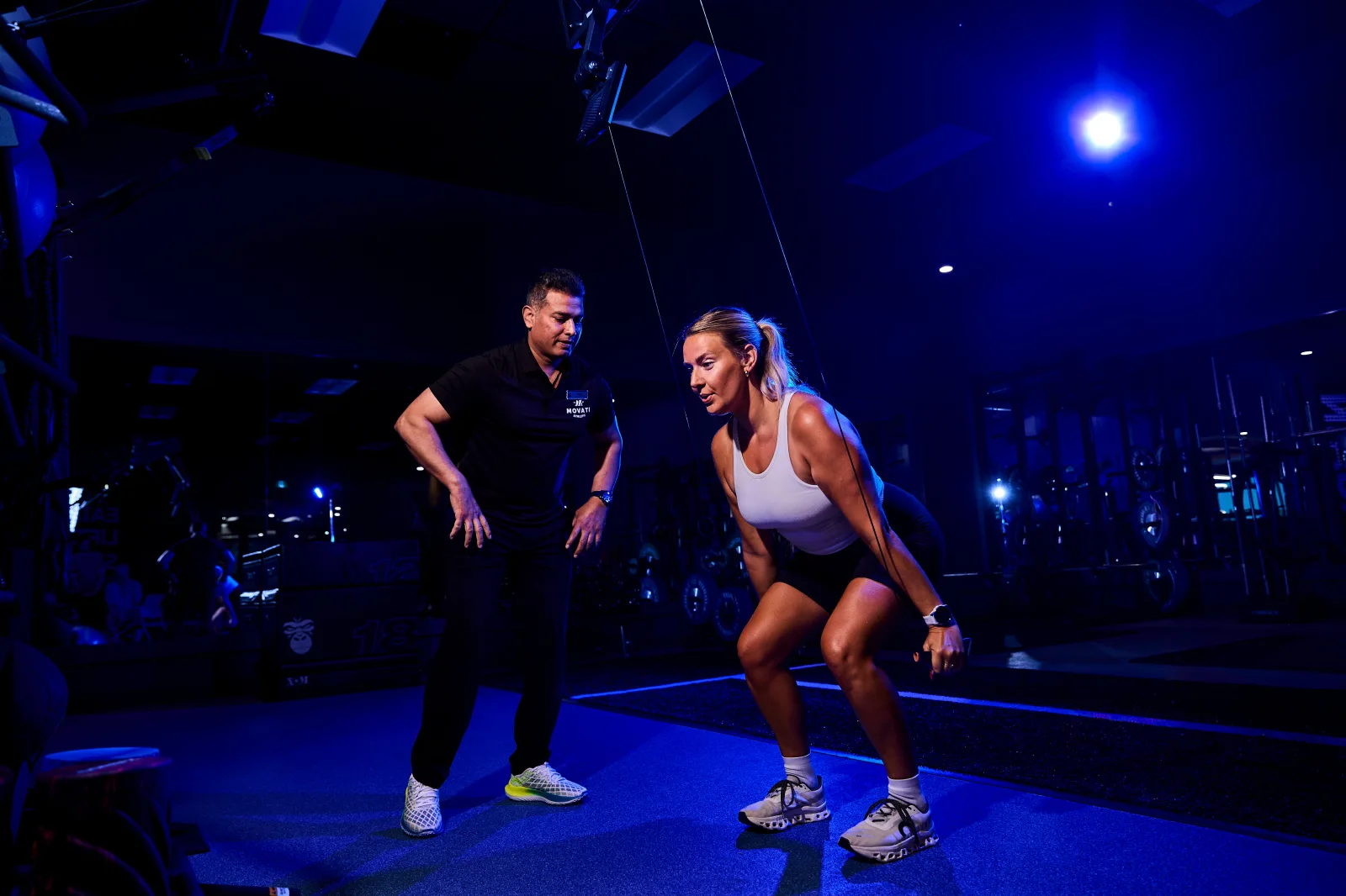 Team MOVATI Takes on HYROX Toronto with a trainer guiding a woman in a gym workout under blue lighting.